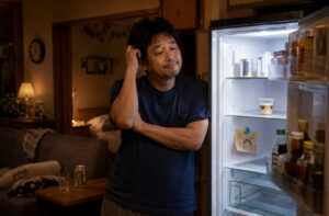 夜の自宅で冷蔵庫を開け、中を見ながらぼんやり考え込む中年男性。